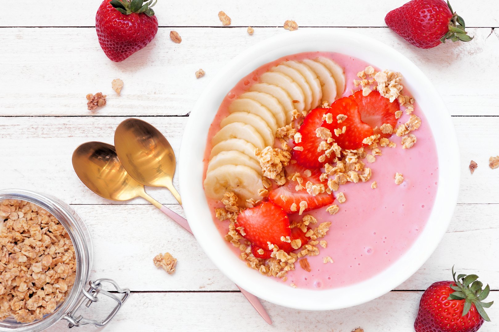 Healthy strawberry and banana smoothie bowl with granola. Above view table scene on a white wood background.