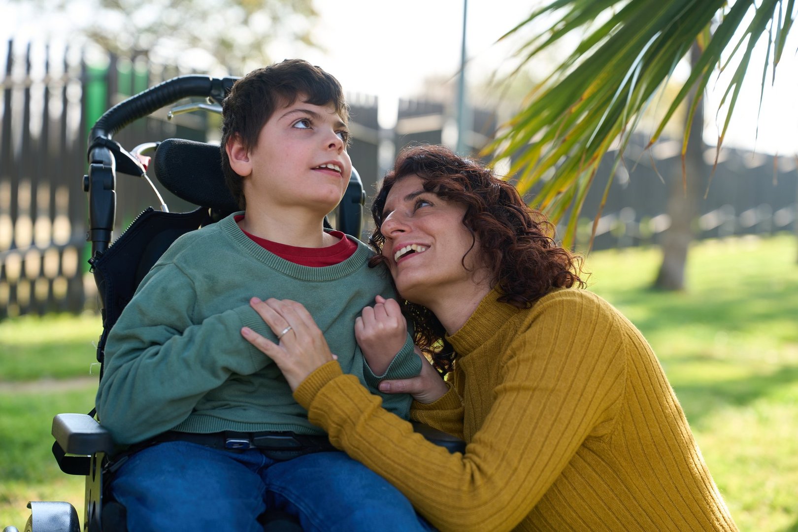 Happy mother hugging her disabled son sitting in a wheelchair, in a park, during a sunny day