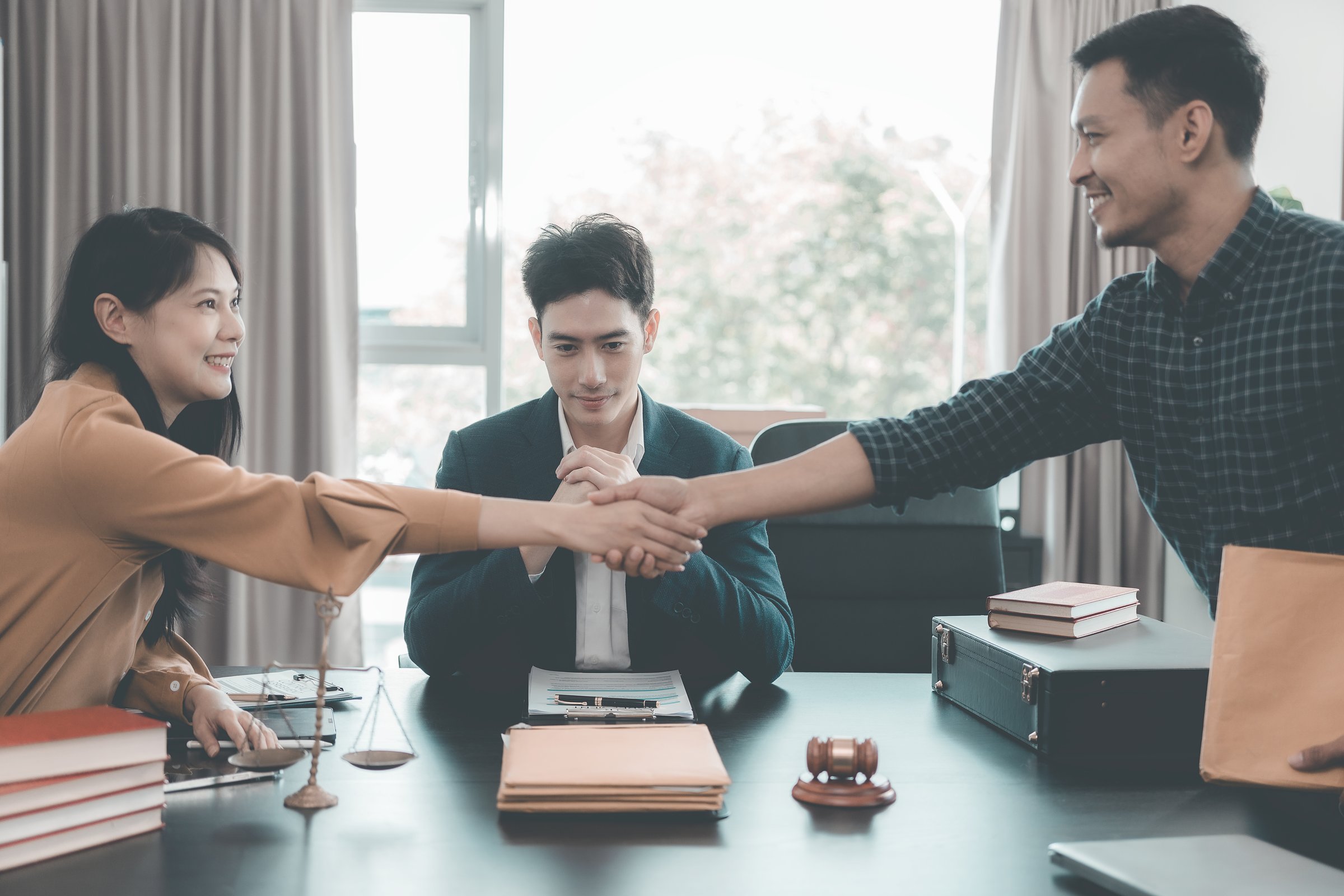 Lawyer shaking hands with client in her office representing legal services and signing agreement with hammer and scales of justice in foreground