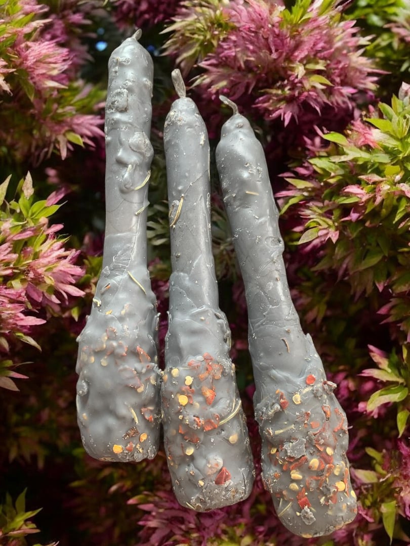 Three wax-dipped vegetable-shaped objects with red chili flakes, hanging in front of purple-leaved plants.