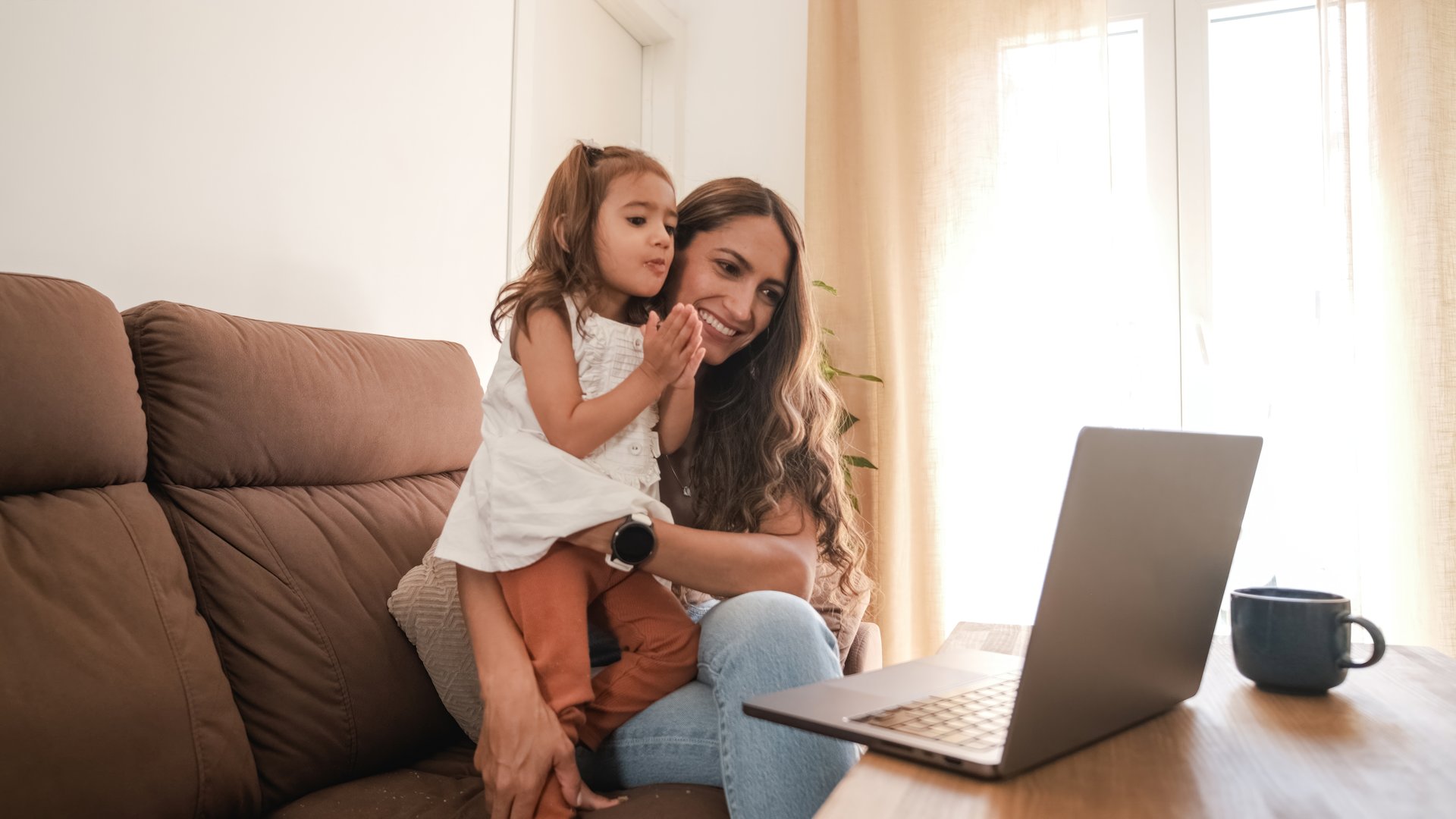 Smiling Latin mother in casual clothes showing laptop to small daughter while watching cartoon on sofa during weekend in cozy living room of apartment