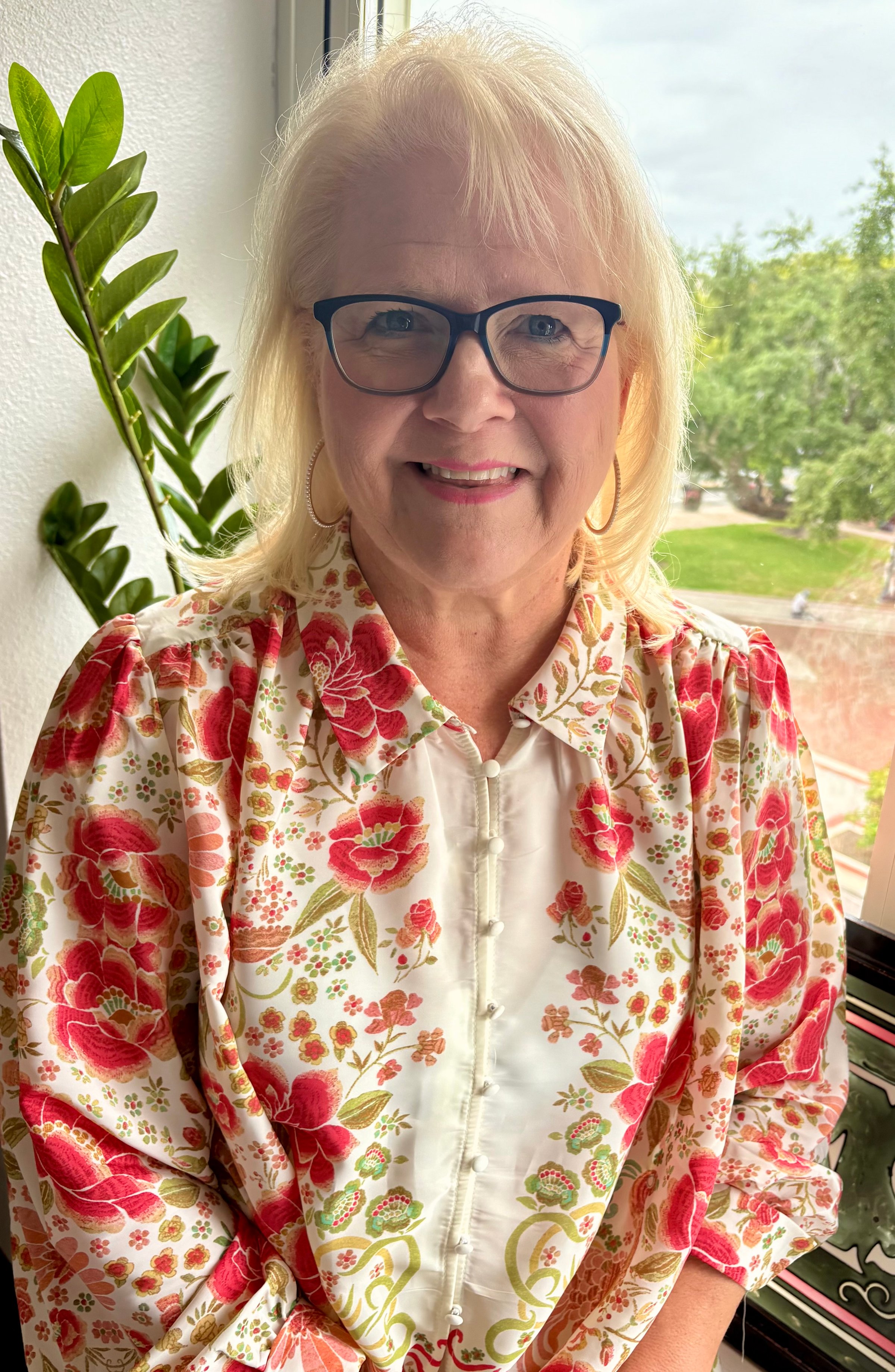 Smiling woman with glasses in floral blouse standing by a window with a plant and trees visible outside.