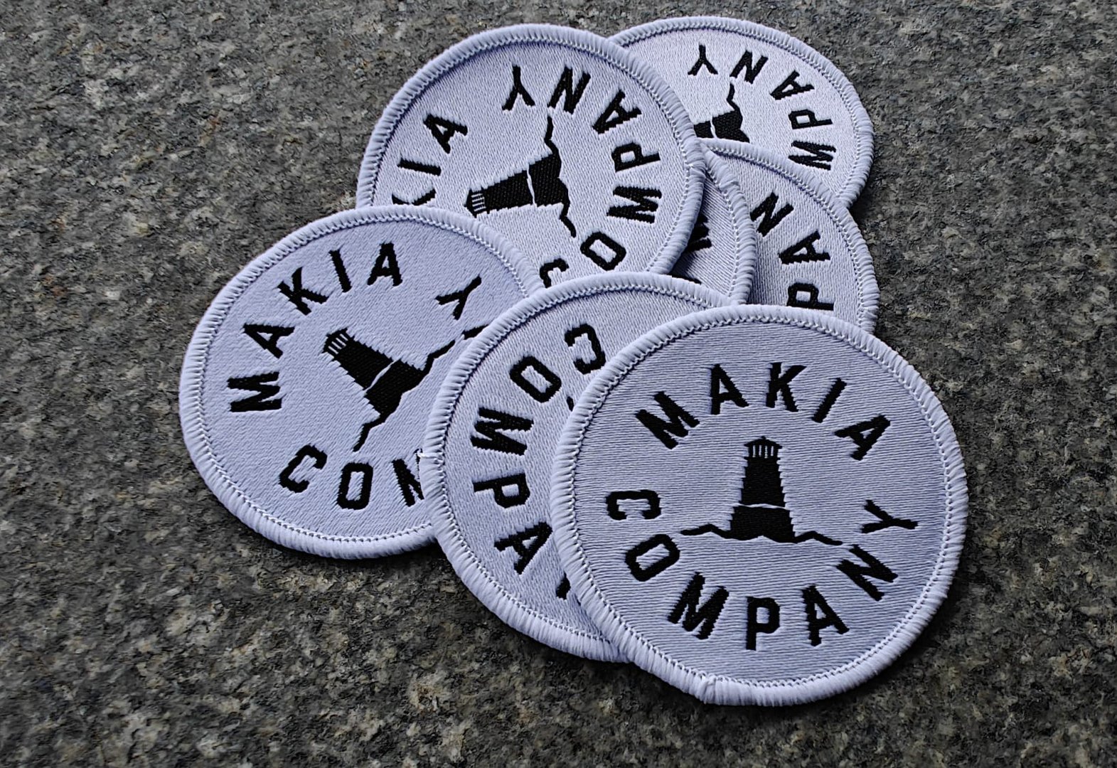 Circular patches featuring a lighthouse design and Makia Company text, arranged on a gray textured surface.
