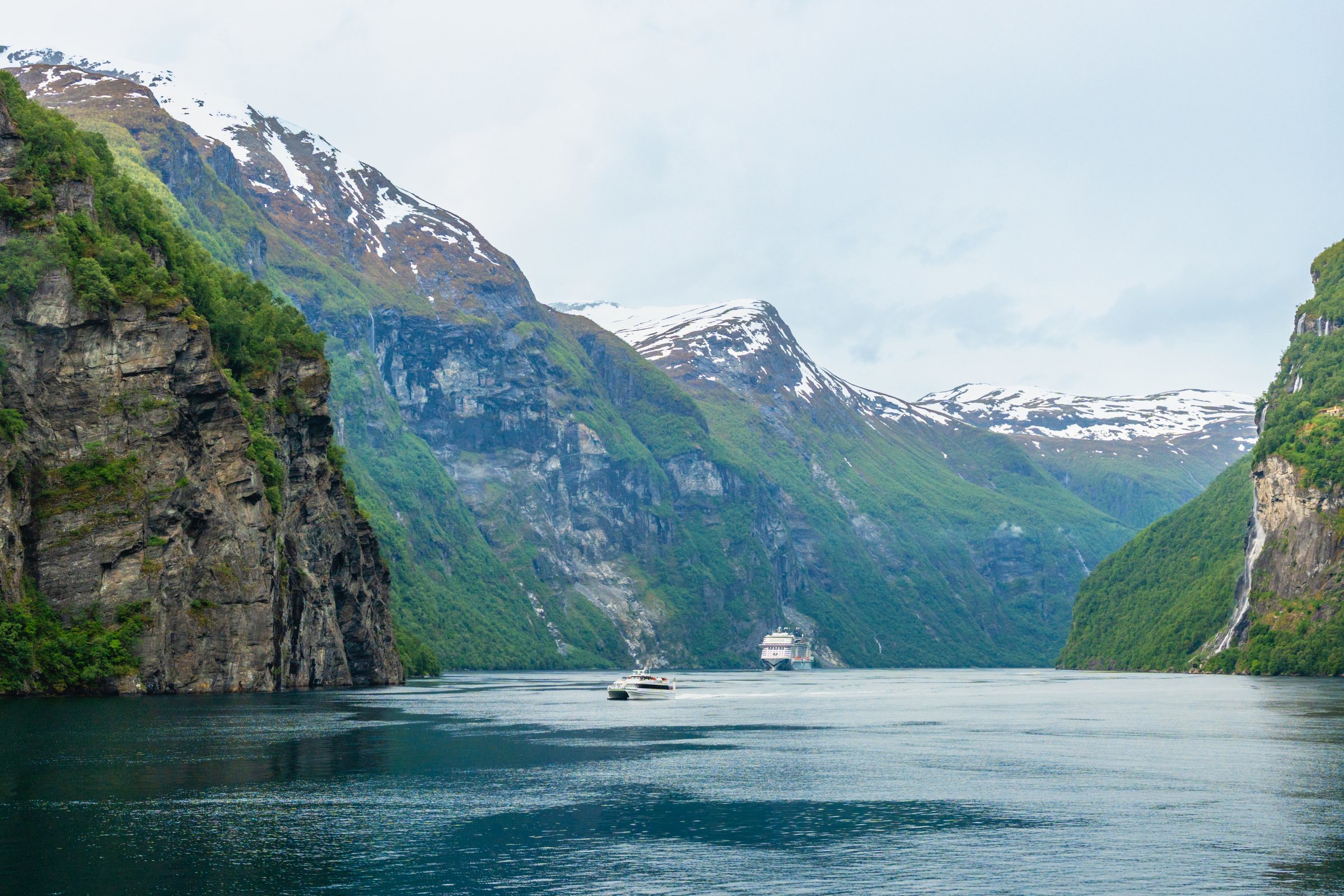 Geirangerfjord, Norway - June 10th 2025:  Lush green foliage on rocky mountain cliffs, snowcapped mountains, and waterfalls, line Norway's most gorgeous UNESCO designated fjord, Geirangerfjord, and dwarf several nautical vessels on the water