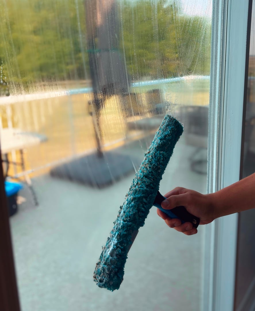 Person cleaning a patio door window with a blue squeegee, sunny patio and pool visible in the background.