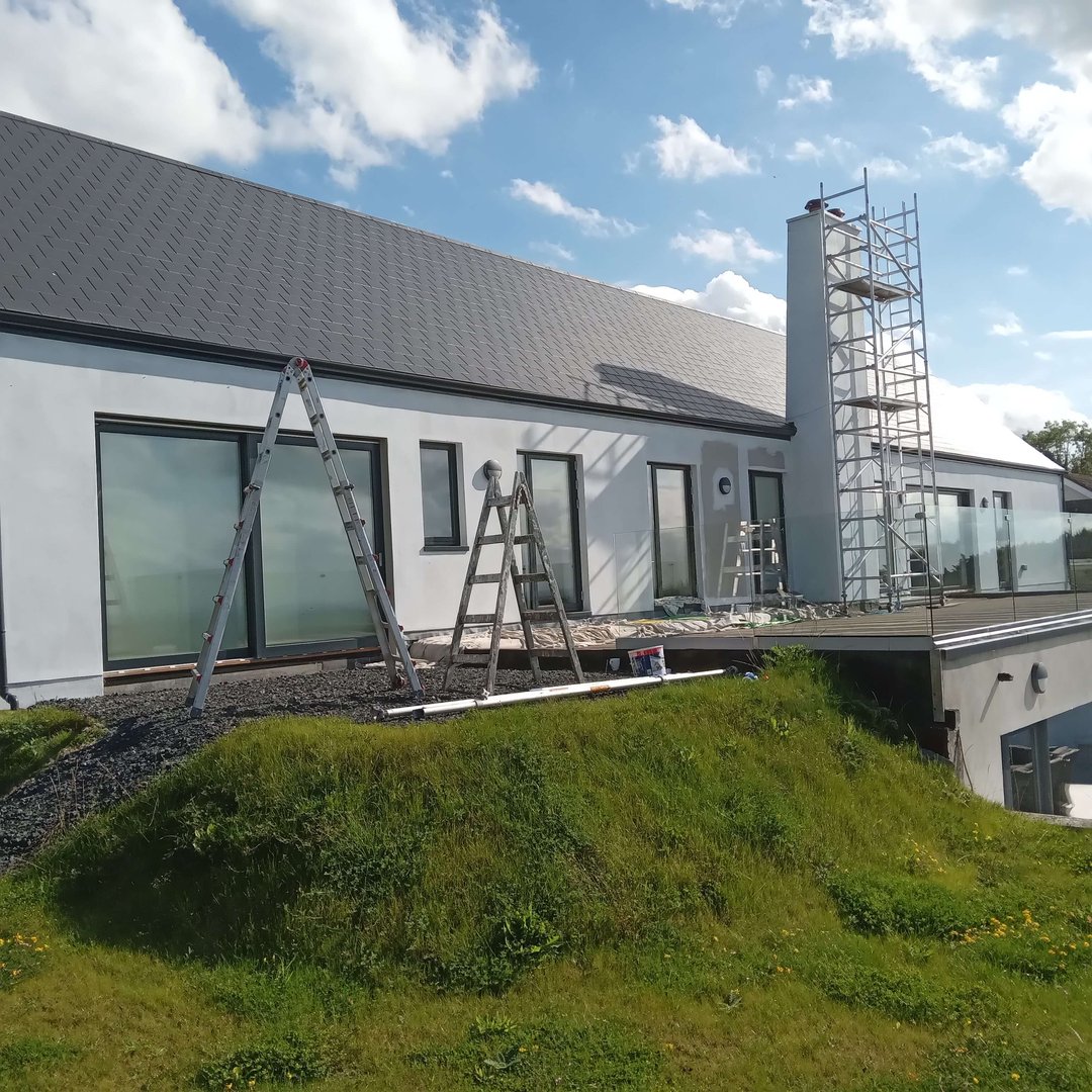 House under construction with ladders and scaffolding, featuring a gray roof and white exterior. Cloudy sky in the background.