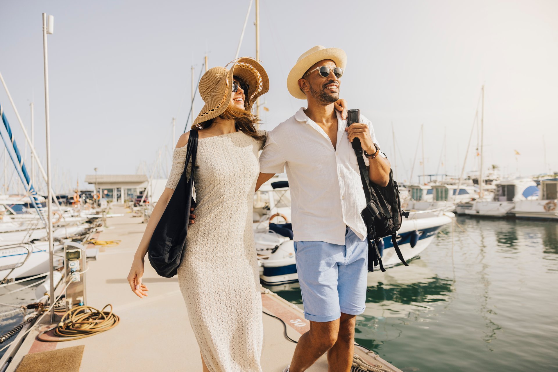 A smiling couple walks hand in hand along the marina's dock, surrounded by boats on a sunny day.