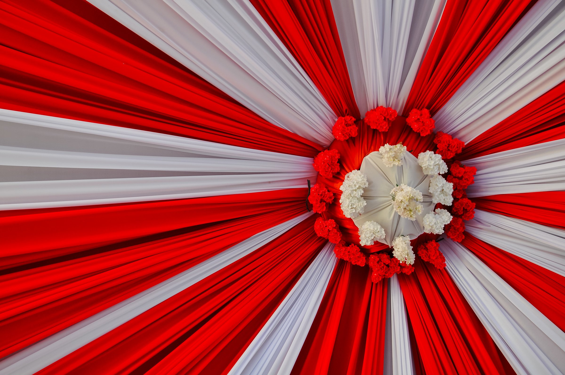 A detailed view of a beautiful ceiling decoration featuring red and white fabric drapes arranged in a radial pattern. The centerpiece is adorned with a cluster of vibrant red and white flowers.