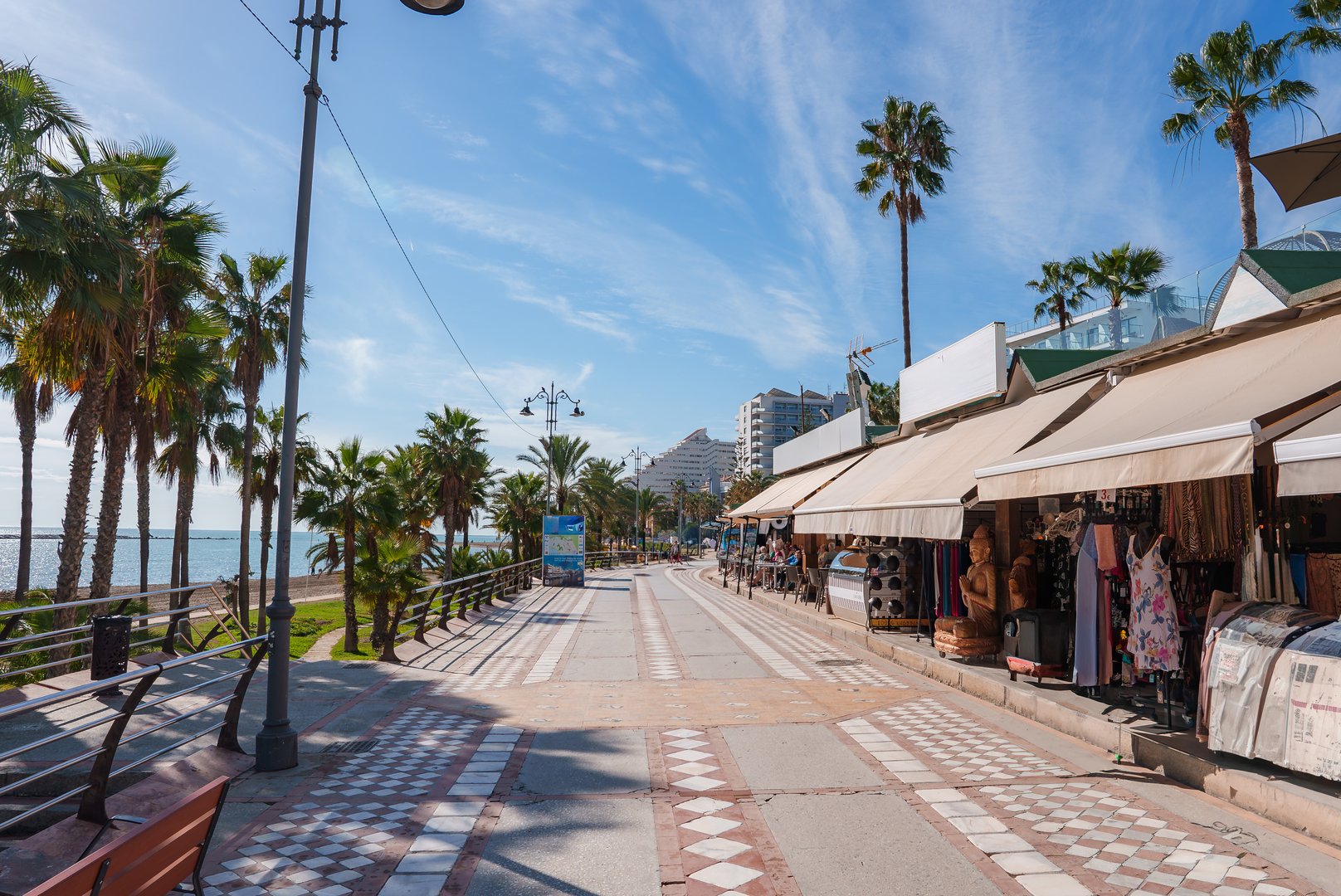 A scenic walkway in Benalmadena, Spain, lined with palm trees and shops. Distinctive tiled path, modern buildings, and sea visible under a clear blue sky.