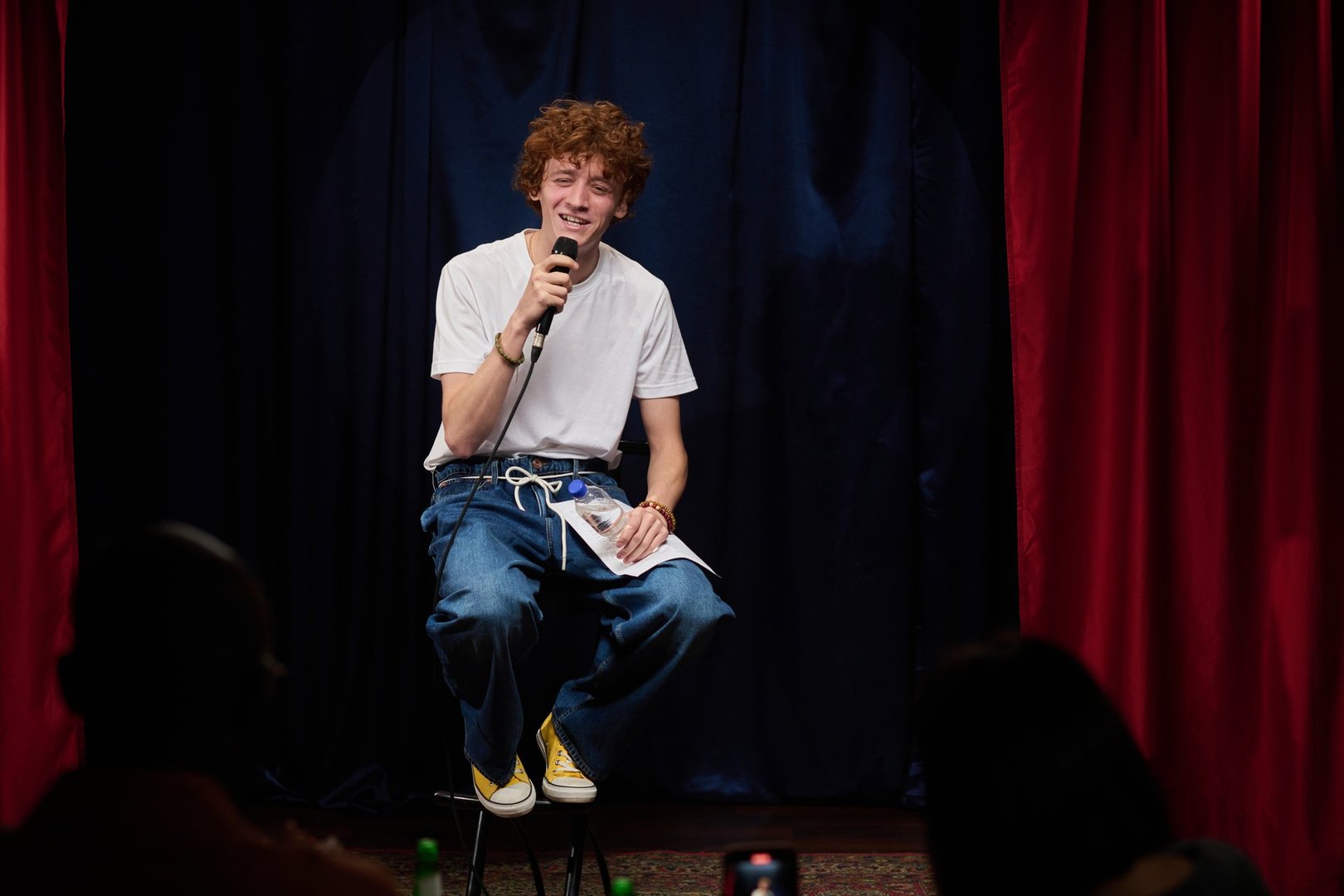 Young performer holding microphone and reading notes while smiling on dark stage during comedy show in front of audience surrounded by red curtains
