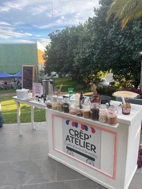 Outdoor crepe stand with various toppings in jars, a menu board, and a coffee dispenser in a park setting.