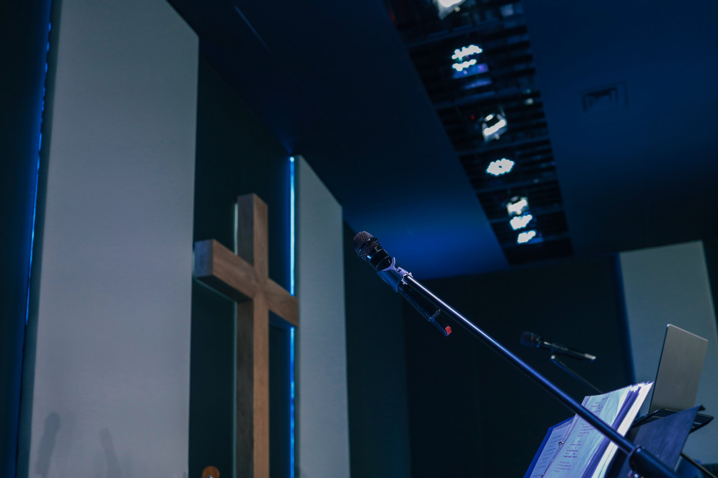 Microphone on a stand with open sheet music and a wooden cross in the background inside a modern worship hall.