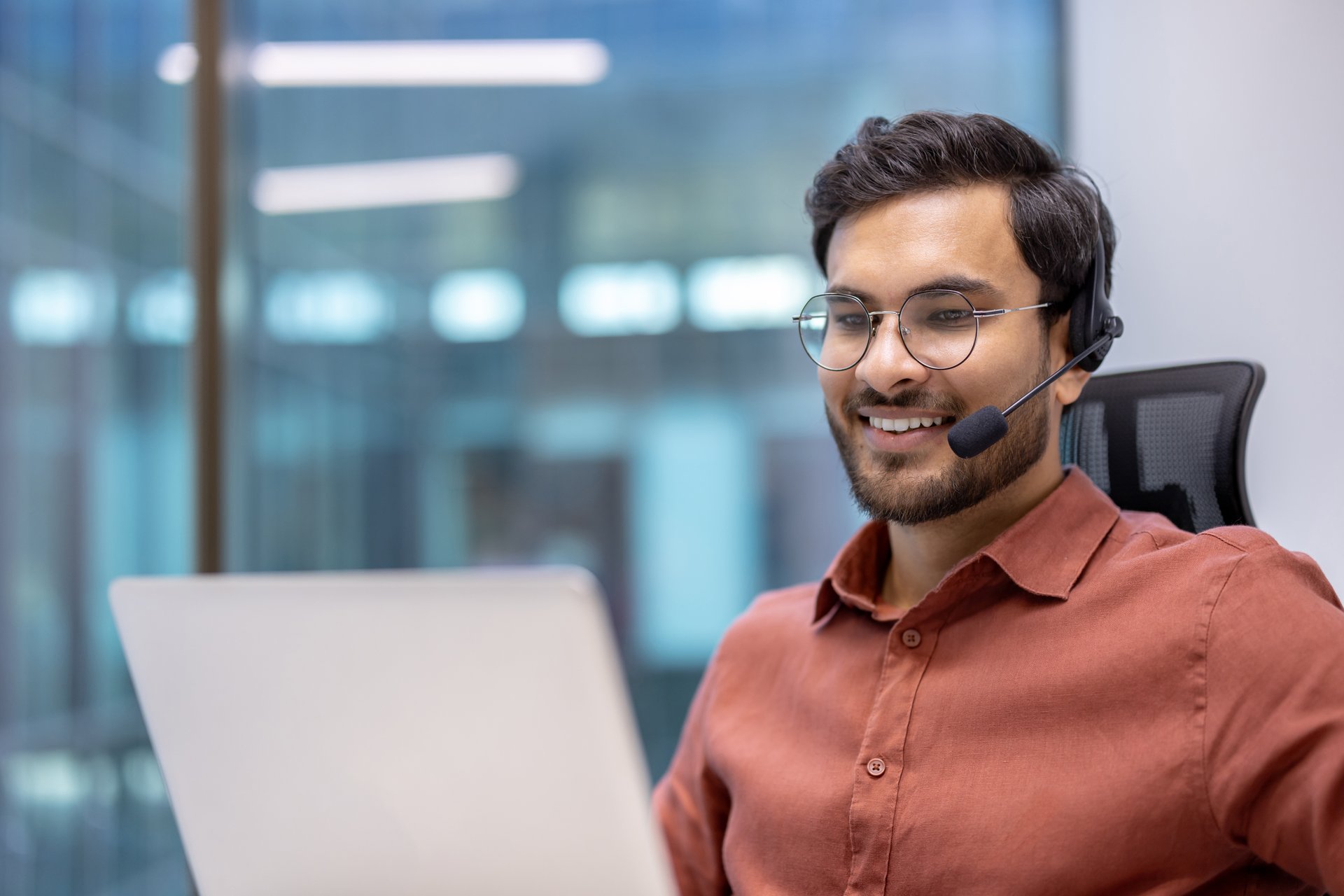 Businessman wearing headset focused on video call. Office environment with large windows. Engaged in communication, showcasing professionalism and teamwork in corporate setting.