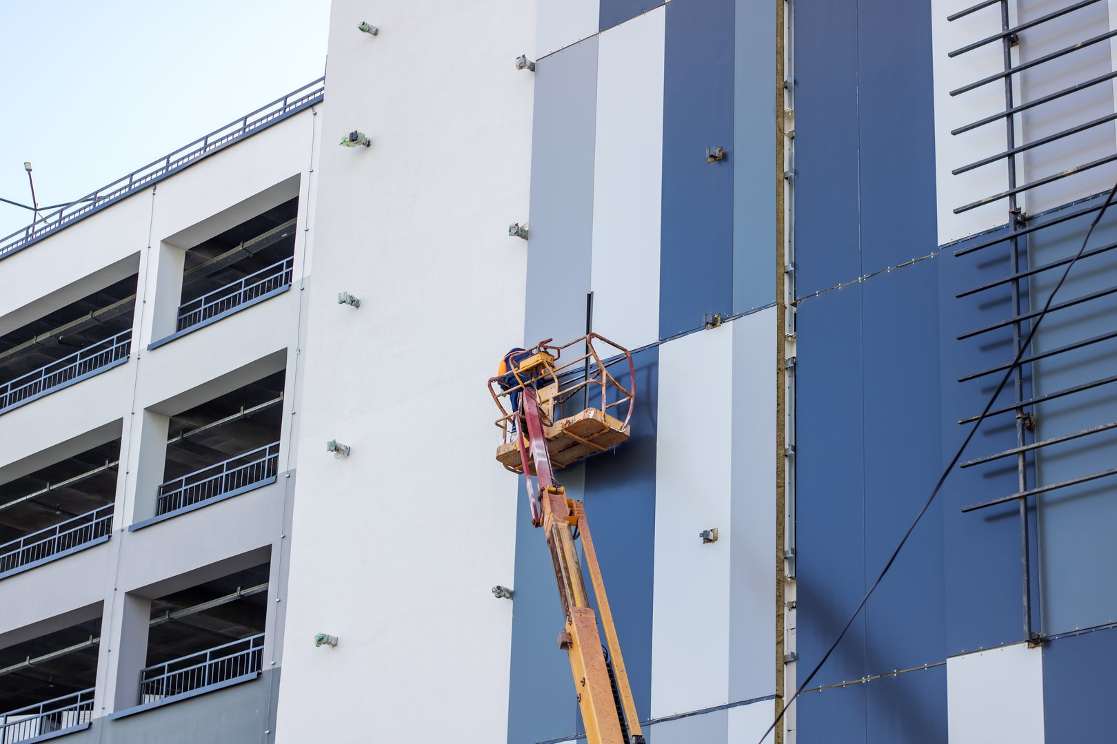 A man is seen inside a lift painting the side of a building. The building is a tower block or condominium in an urban setting. A crane and electricity lines can be spotted nearby