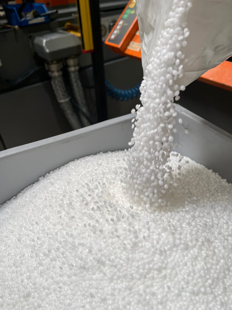 pouring of a bag of white plastic pellets on a gray plastic drawer, production of plastic parts, plastic injection, spilling of plastic pelets, with a plastic injector out of focus in the background, small plastic granules vertical