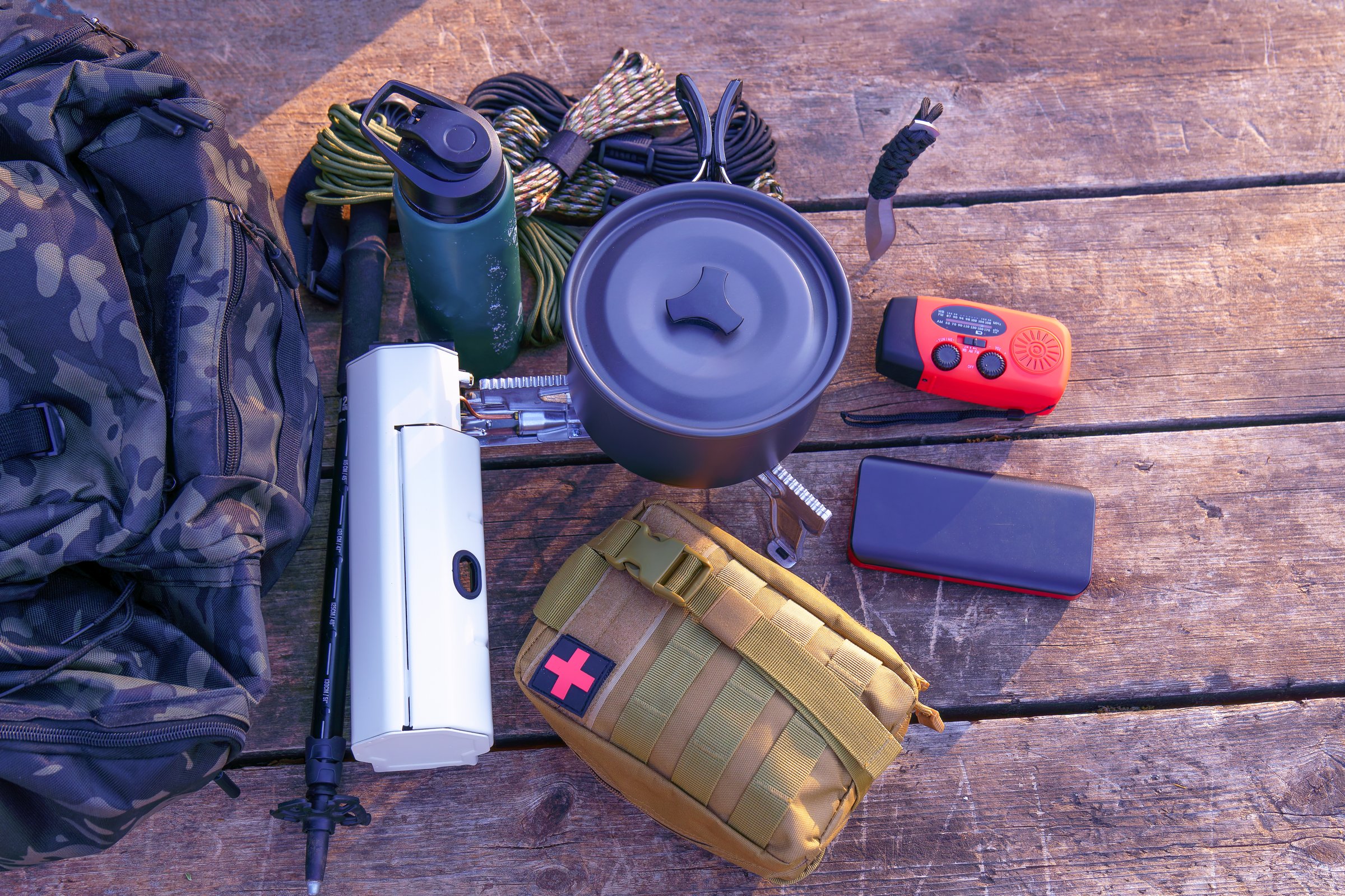 Essential camping gear including backpack, cooking pot, first aid kit, and navigation tools arranged on a weathered wooden surface