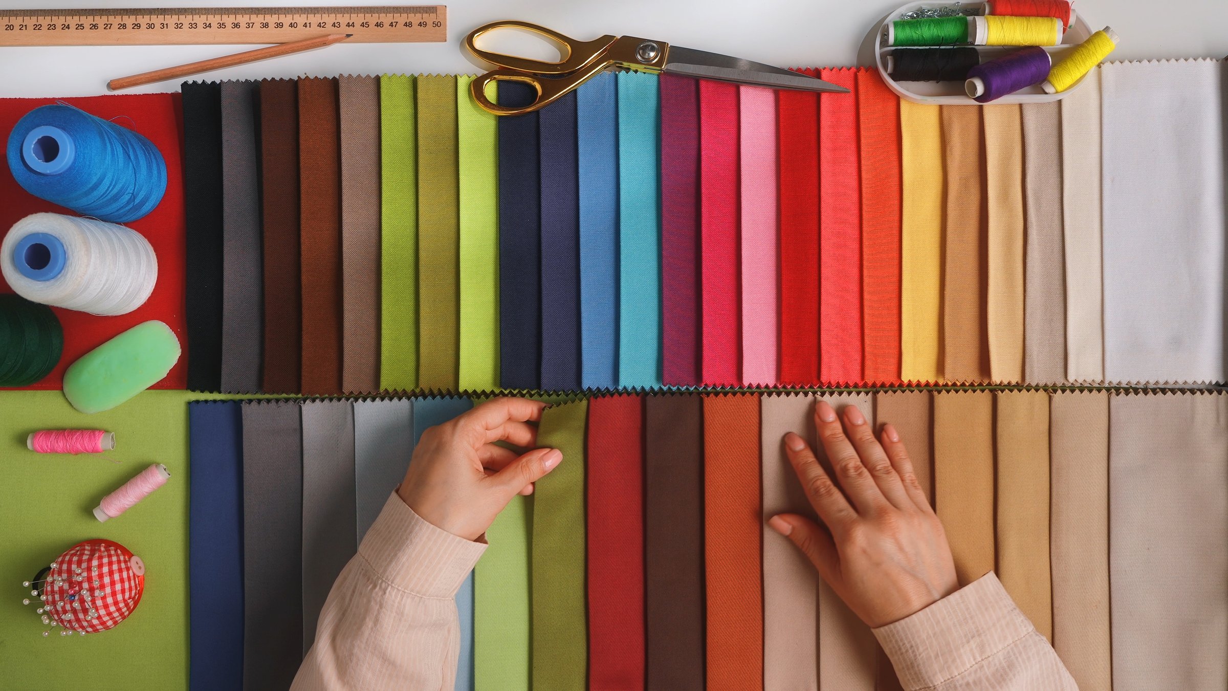 Top view of a designers hands examining fabric swatches. A vibrant collection of sewing supplies including fabric, thread, scissors and ruler, perfect for needlework.