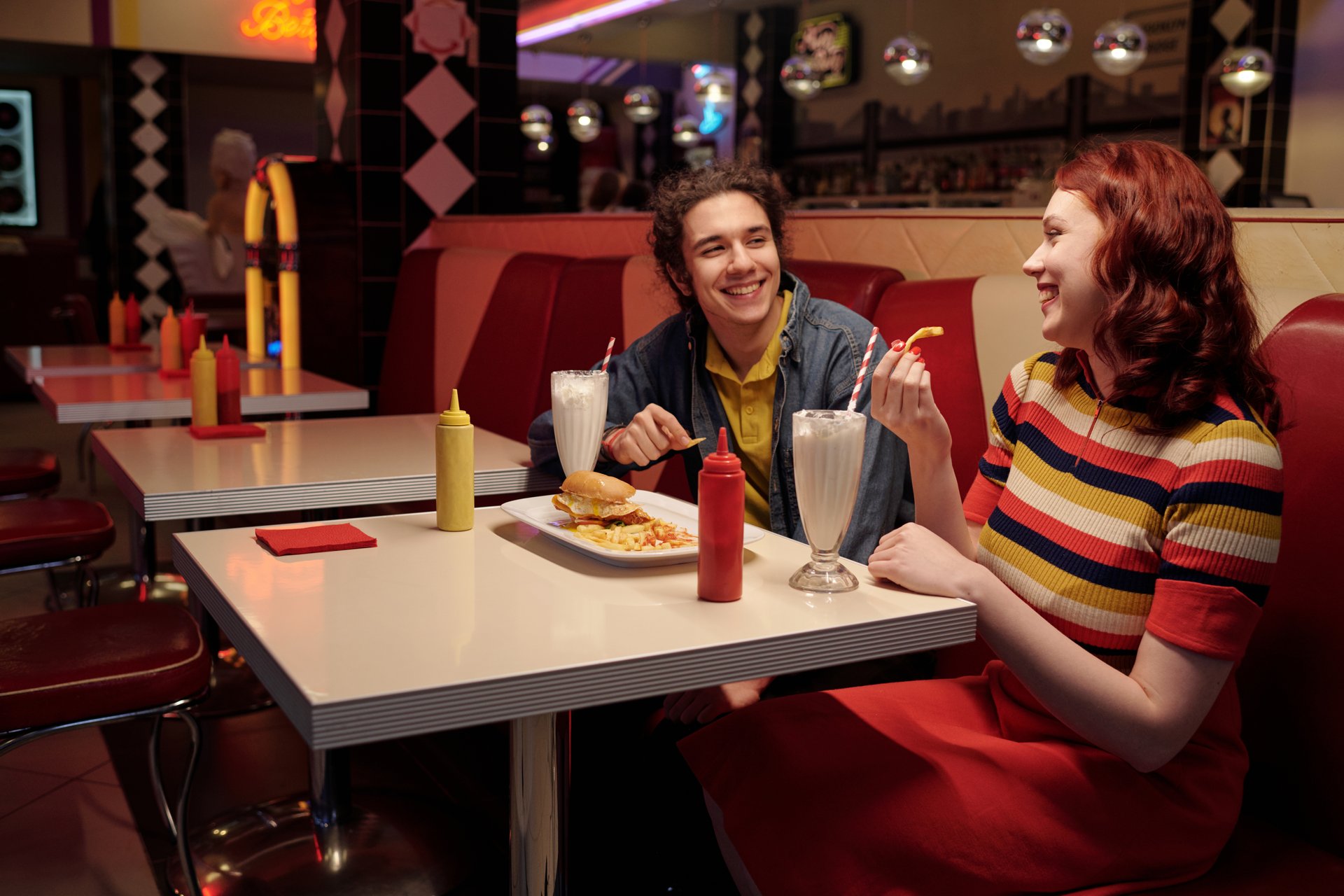 Two friends are enjoying milkshakes and burgers in a classic American diner with red seating booths, decorative elements, and soft lighting, creating a nostalgic atmosphere