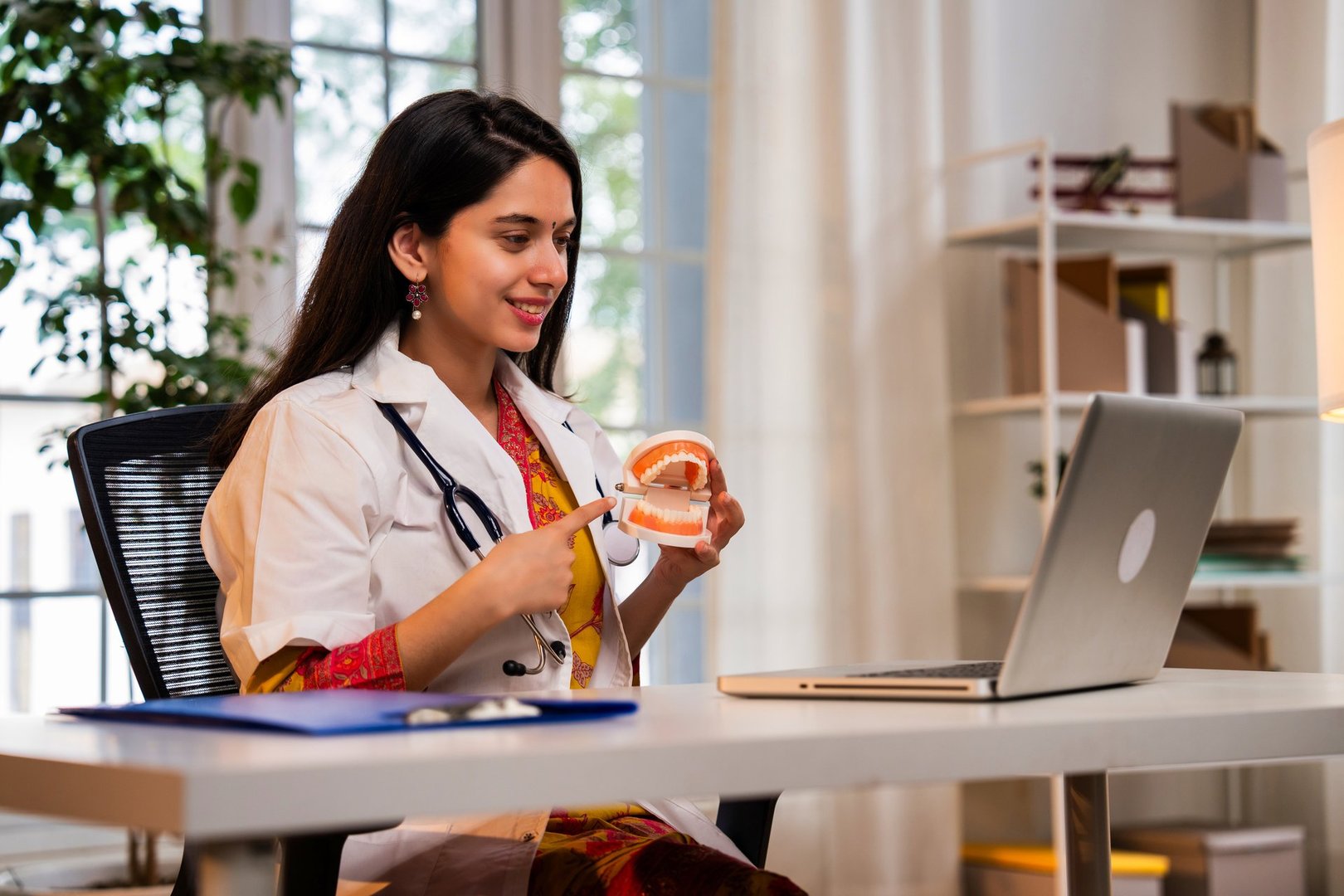 Indian Asian young female dentist wearing a white coat, sitting at a desk with a laptop, explaining oral health care using a model denture during a video call