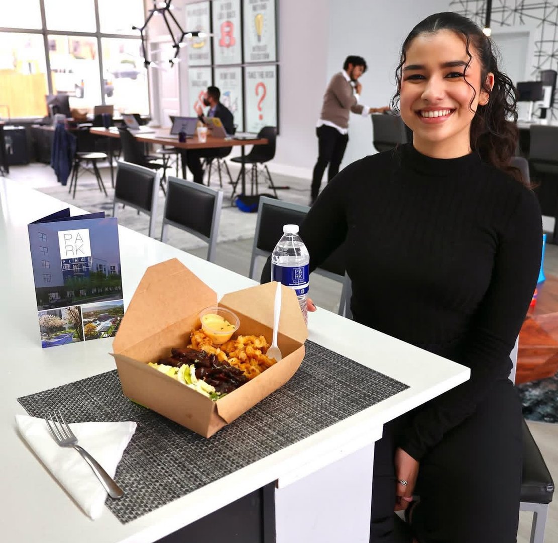 Smiling woman in black sits at a table with a takeout meal and water bottle in a modern cafe.