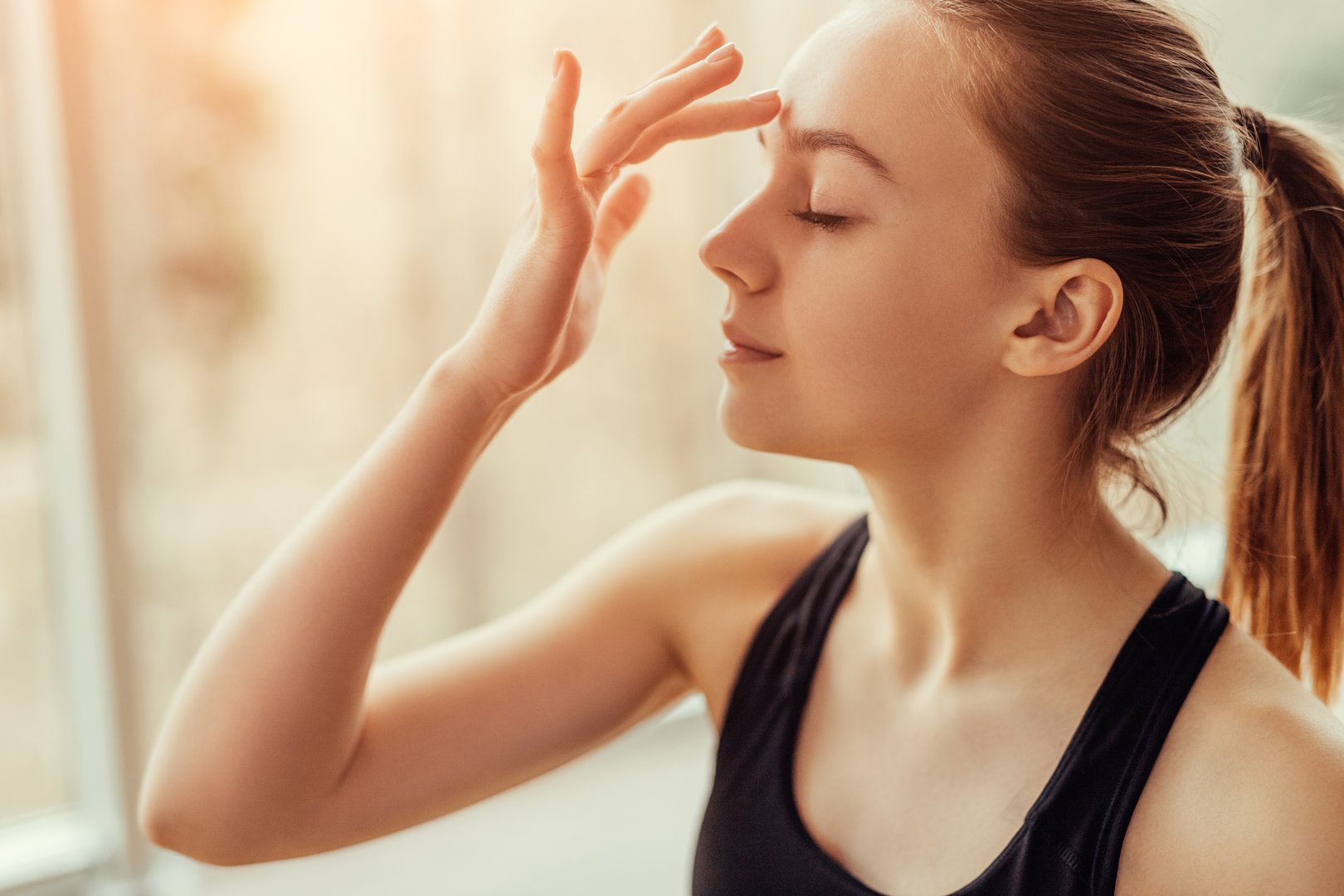 Side view of beautiful young female with closed eyes touching ajna chakra on forehead while doing yoga in gym