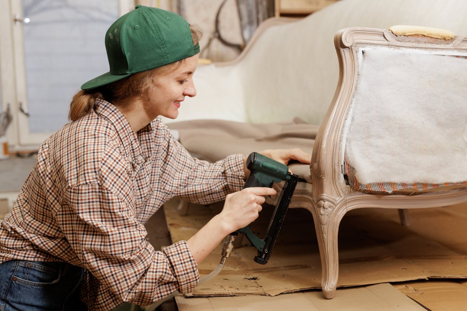 Smiling female upholsterer using a pneumatic staple gun while restoring a vintage armchair in a workshop.