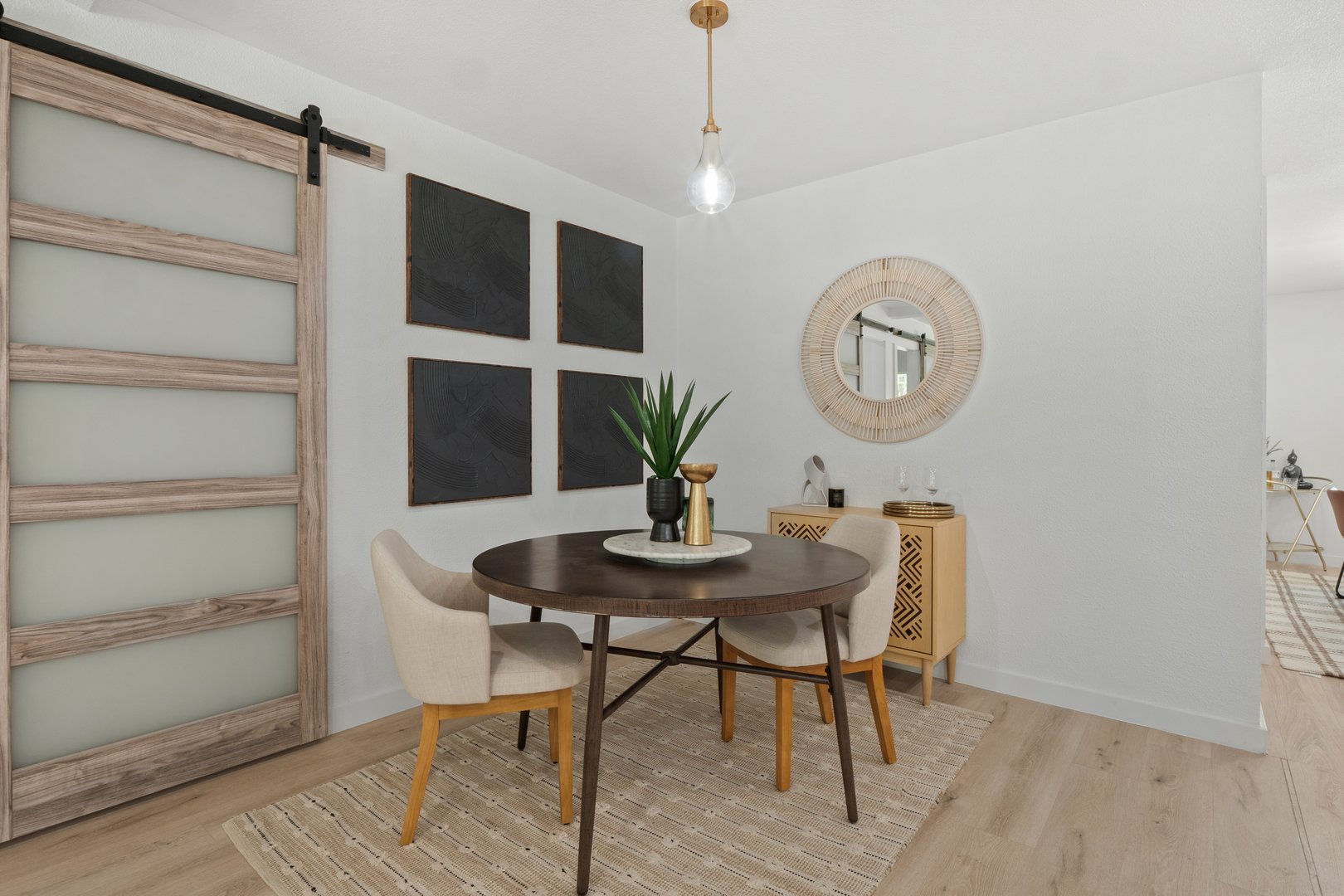 Modern dining area with round table, two chairs, wall art, mirror, and a potted plant on a rug.