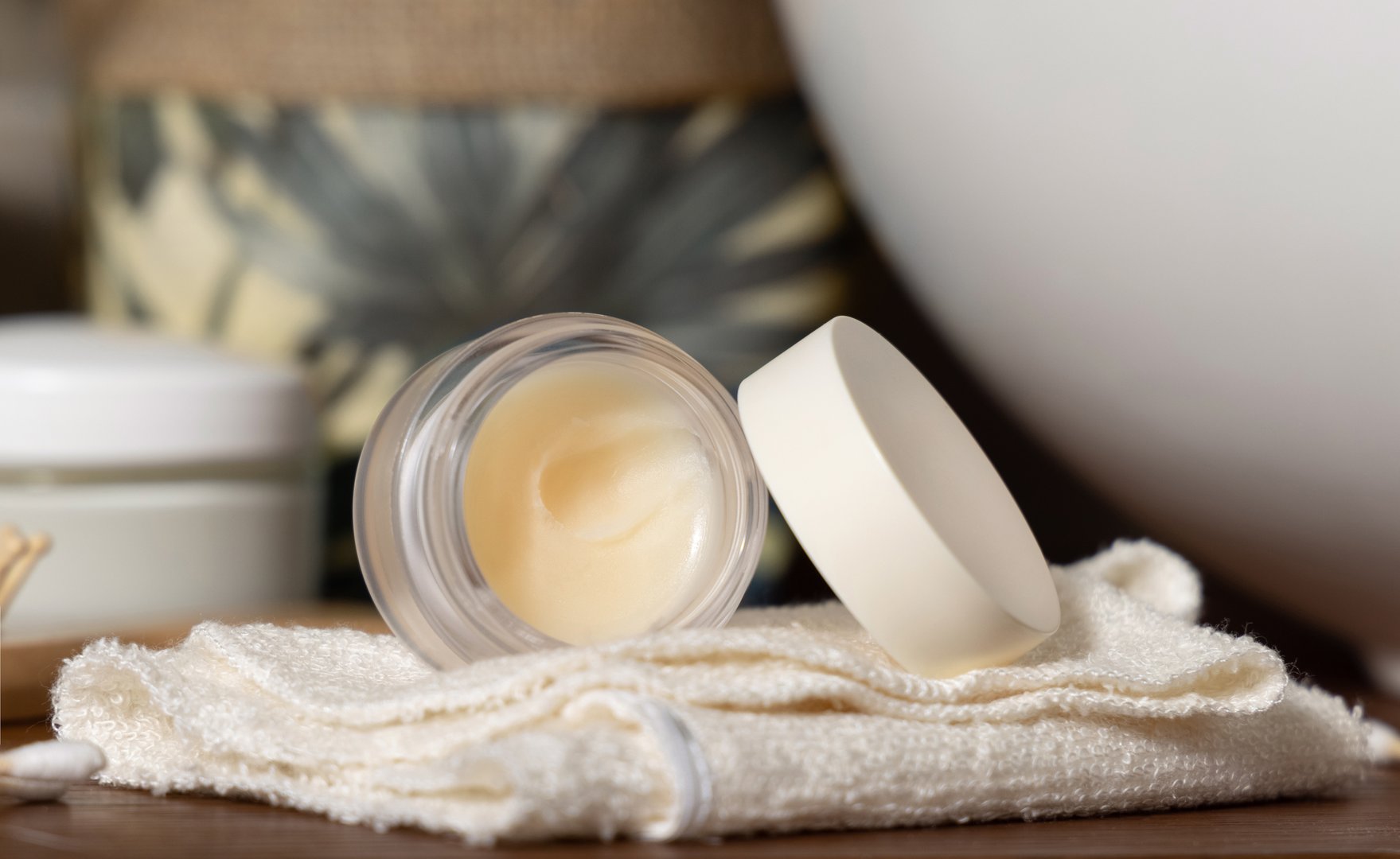 Opened lip balm jar with blank lid on cream folded towel near potted plant and basin on wooden bathroom countertop, close up, cosmetic brand packaging mockup