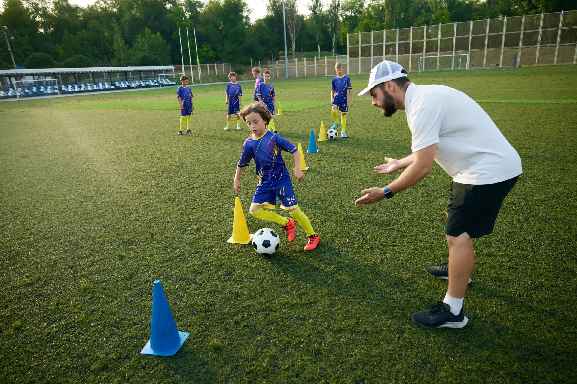 Boy practices dribbling through cones with focused encouragement from the coach. Outdoor stadium and evening game. Concept of sport, childhood, education, achievement, active lifestyle