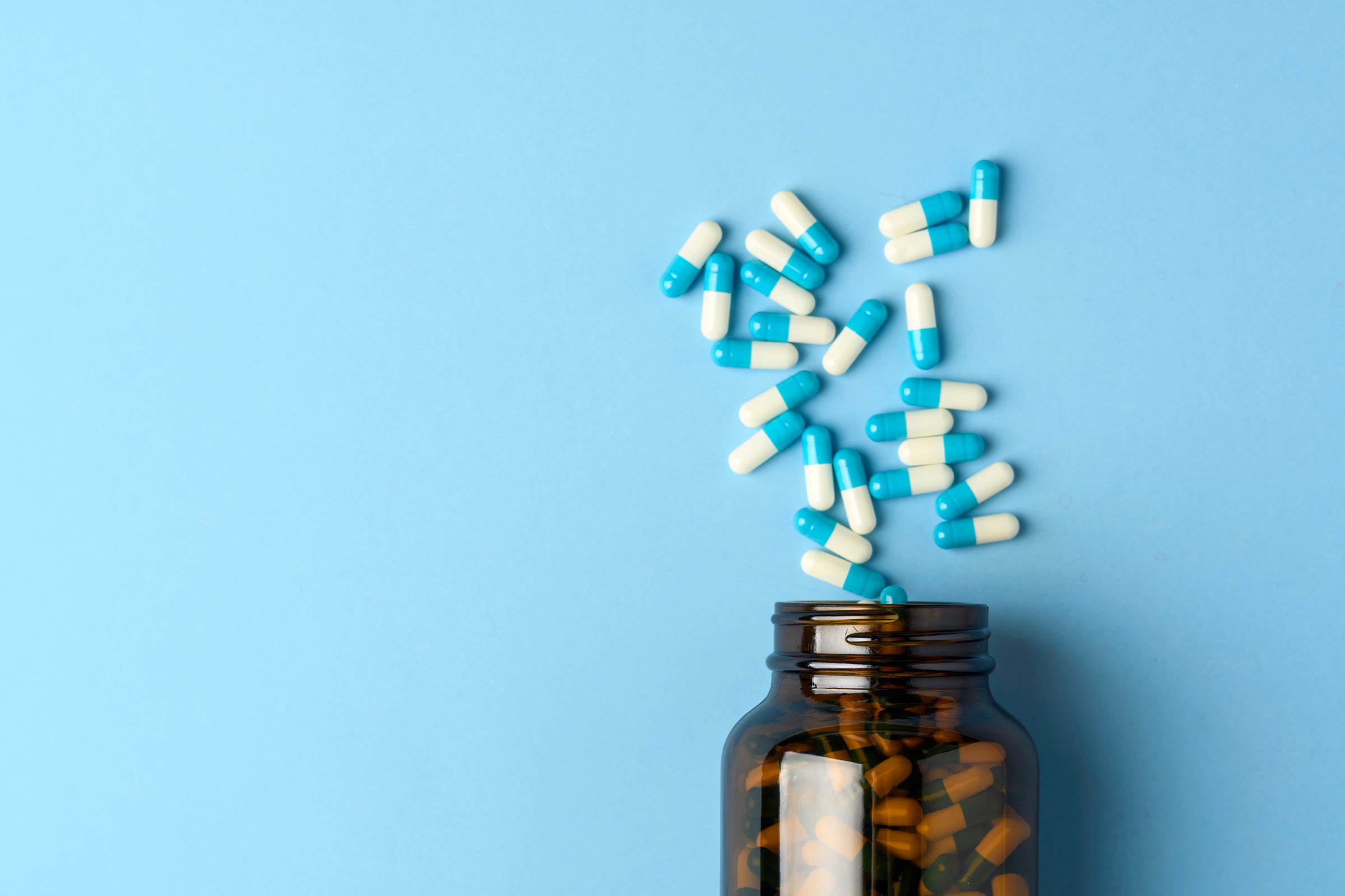 Blue and white capsules spill from amber supplement bottle on pastel blue background with copy space for healthcare ads.