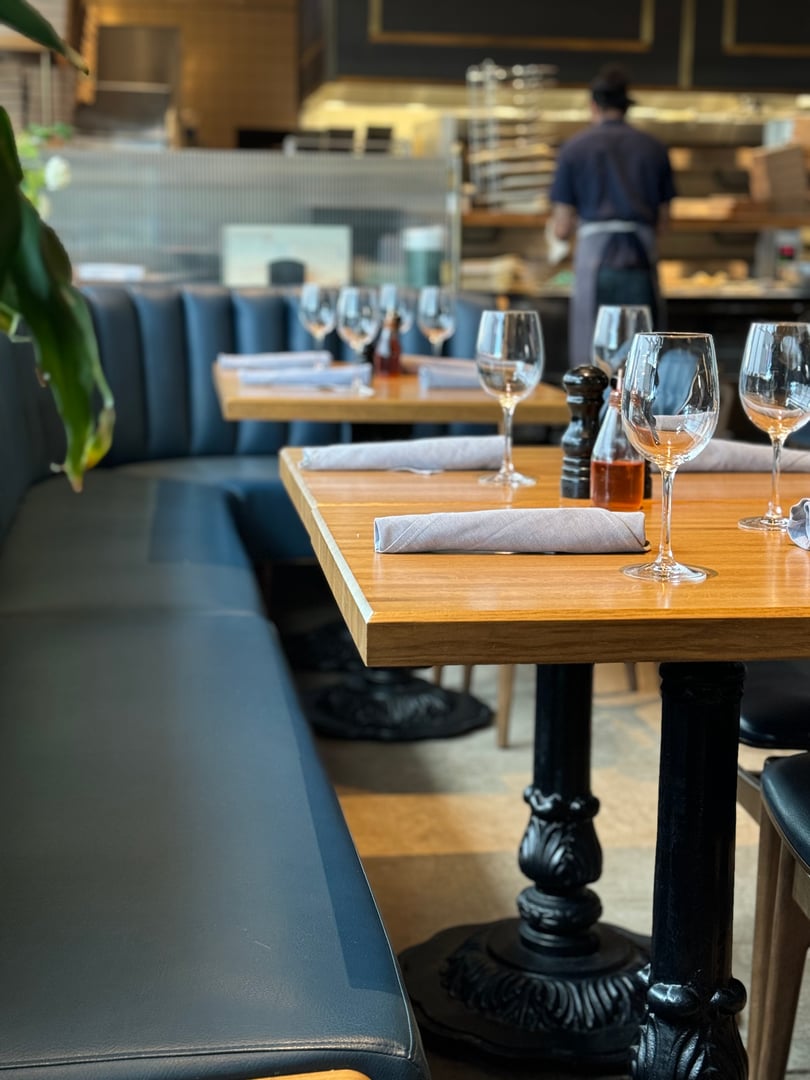 Interior of nice restaurant with empty set tables with black iron stands, blue booth seats and empty wine glasses with blurred back of waiter and kitchen in background.