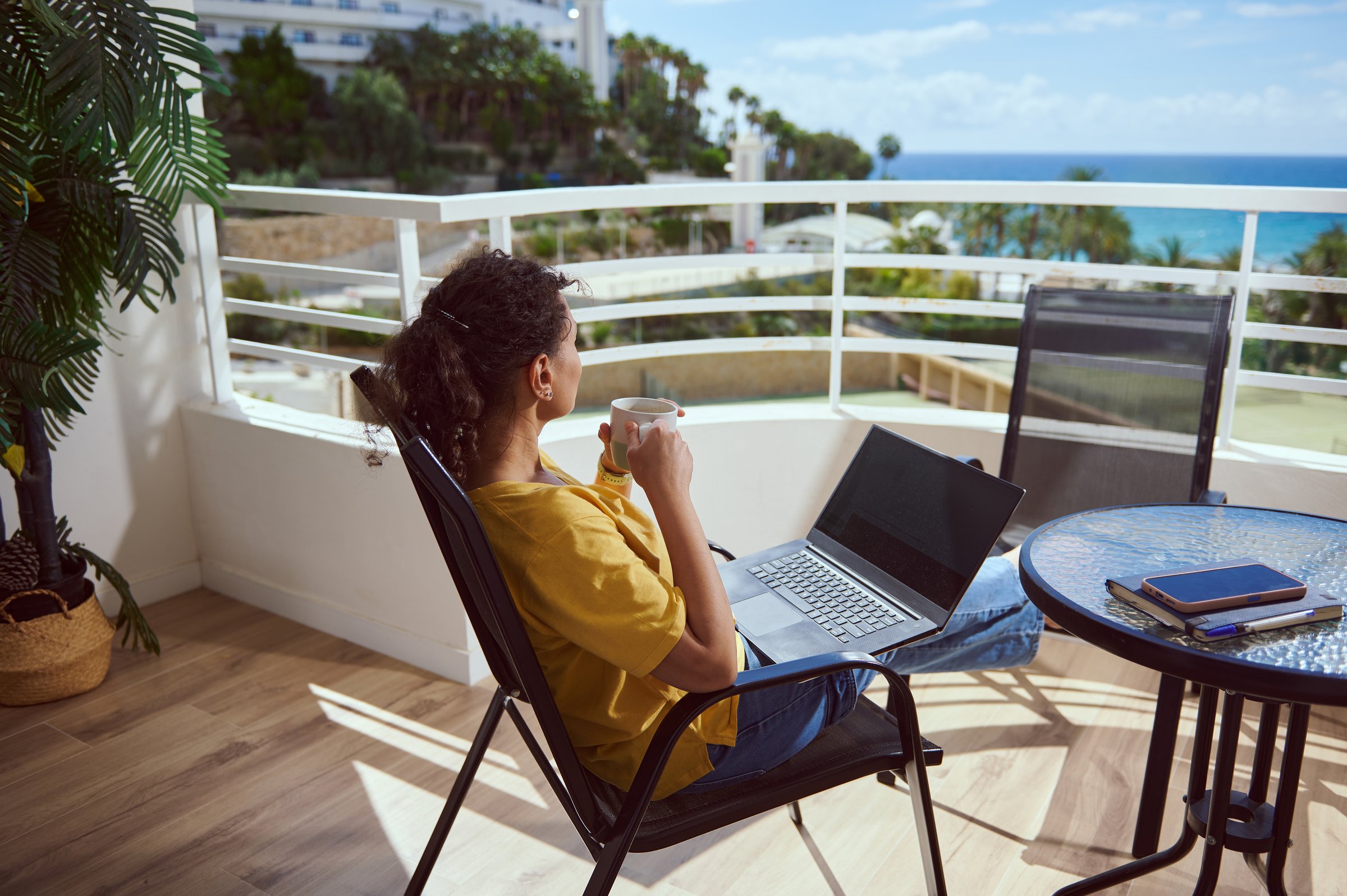 A serene workspace featuring a woman enjoying a drink while using a laptop on a balcony with breathtaking ocean views, showcasing relaxation and productivity.