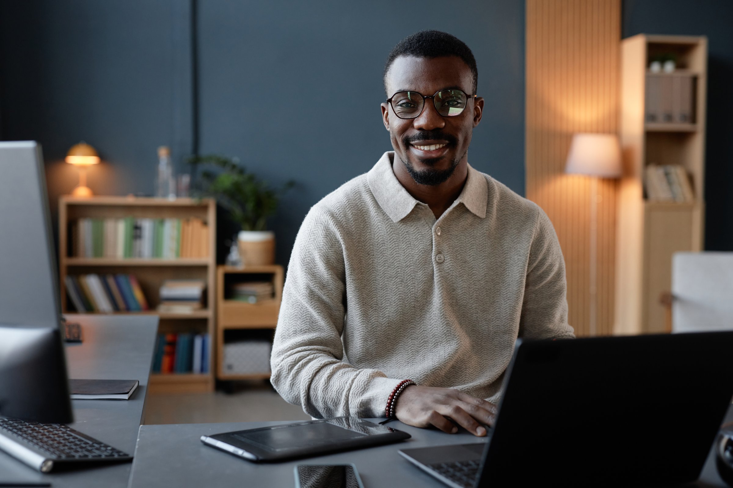 Portrait of cheerful Black male designer with glasses wearing beige polo sweatshirt posing at desk cluttered with devices for digital art in office
