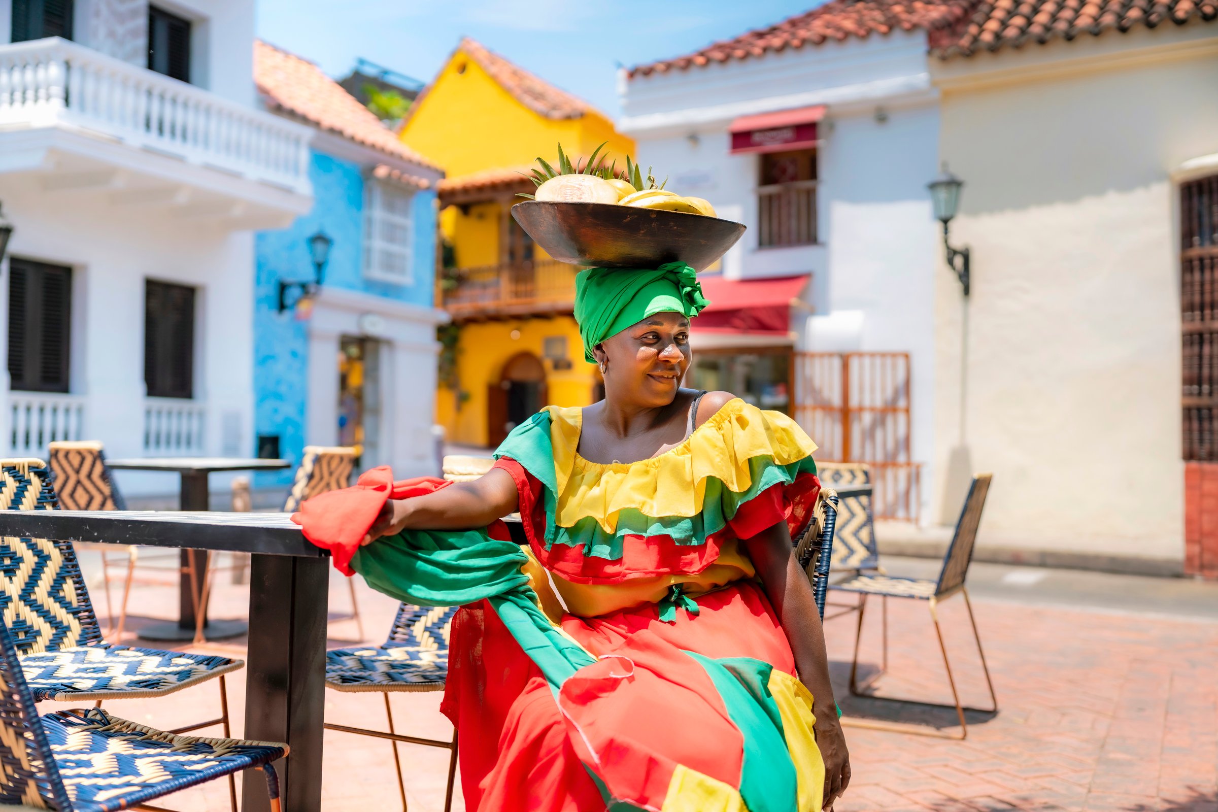 Afro-Latina Palenquera woman in colorful red, green, and yellow outfit sitting with fruit bowl on head in Cartagena, Colombia. Palenqueras are cultural icons of Afro-Colombian heritage.