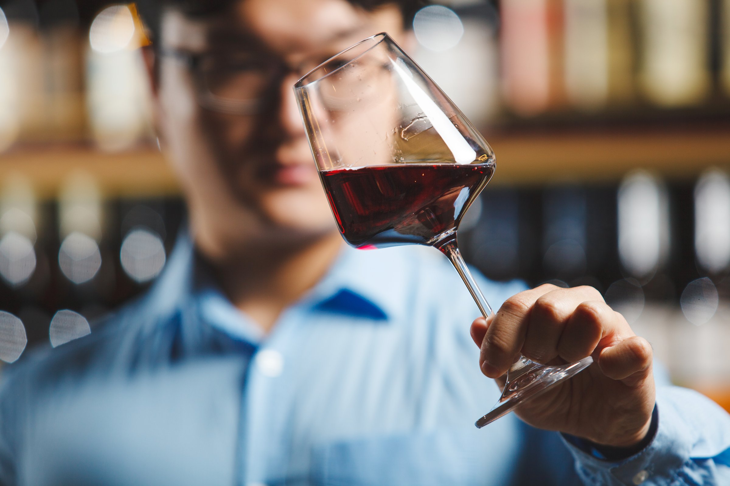 Person closely inspects color and clarity of red wine held in clear stem glass, assessing its quality through visual examination against softly blurred background, being in winery or cellar of wine shop.