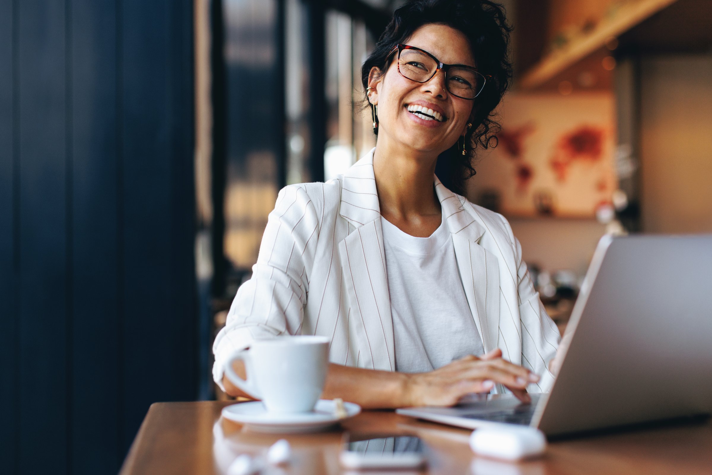 Happy businesswoman working remotely on her laptop in a cozy cafe. Energized freelance worker using smartphone and enjoying her coffee.