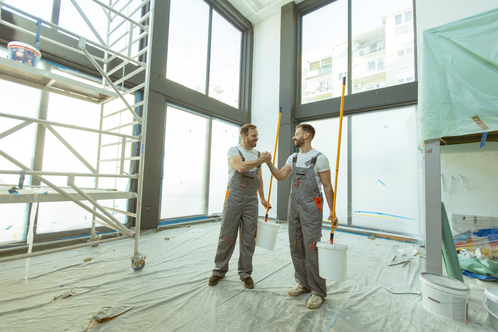 Two workers show support and teamwork while holding paint buckets in a new room with large windows. The room has scaffolding, and the floor is covered with a tarp.