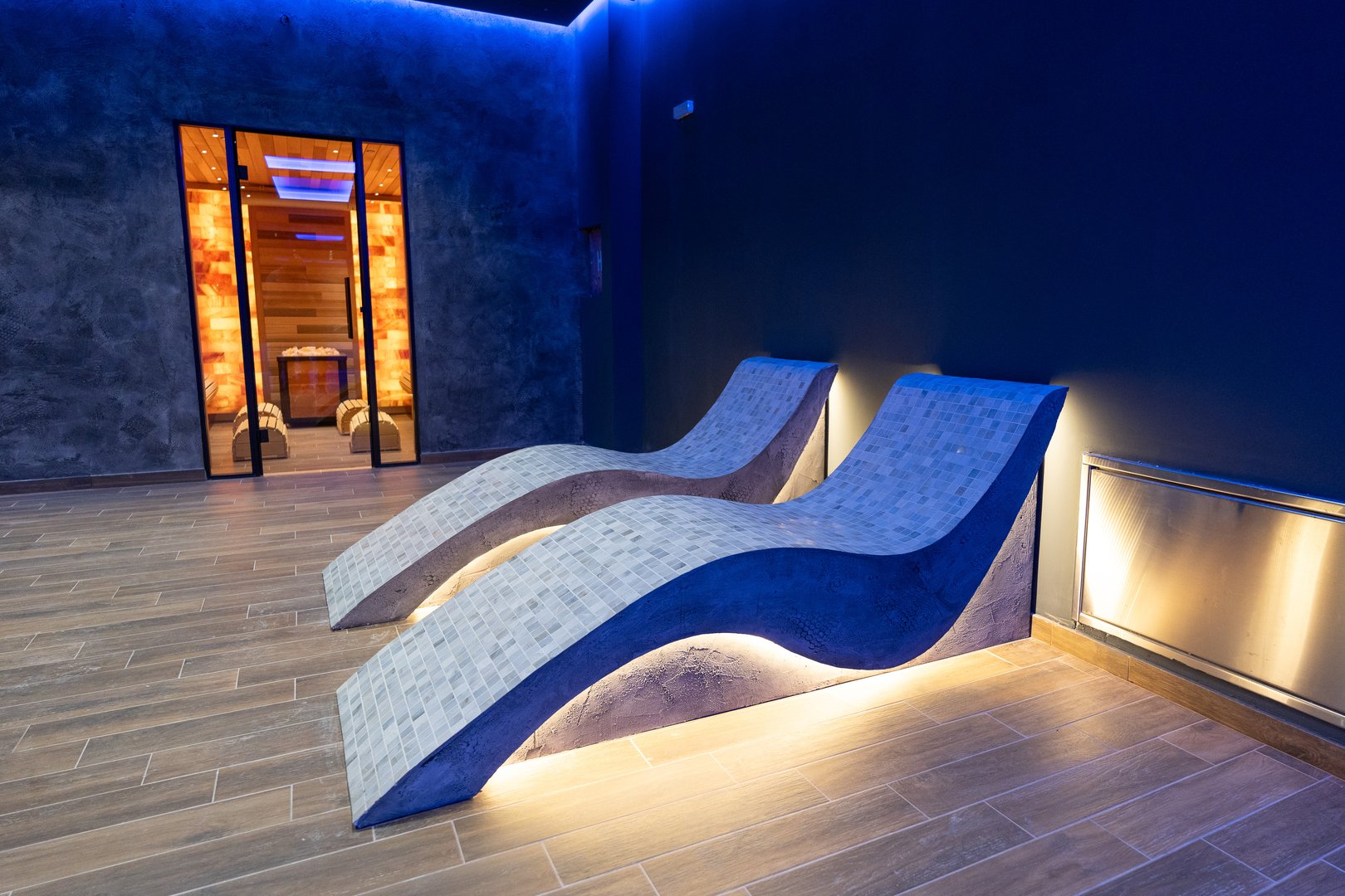 Modern wellness space showcasing elegant heated loungers and sauna entrance, illuminated by soothing ambient blue lighting, evoking calm and relaxation.
