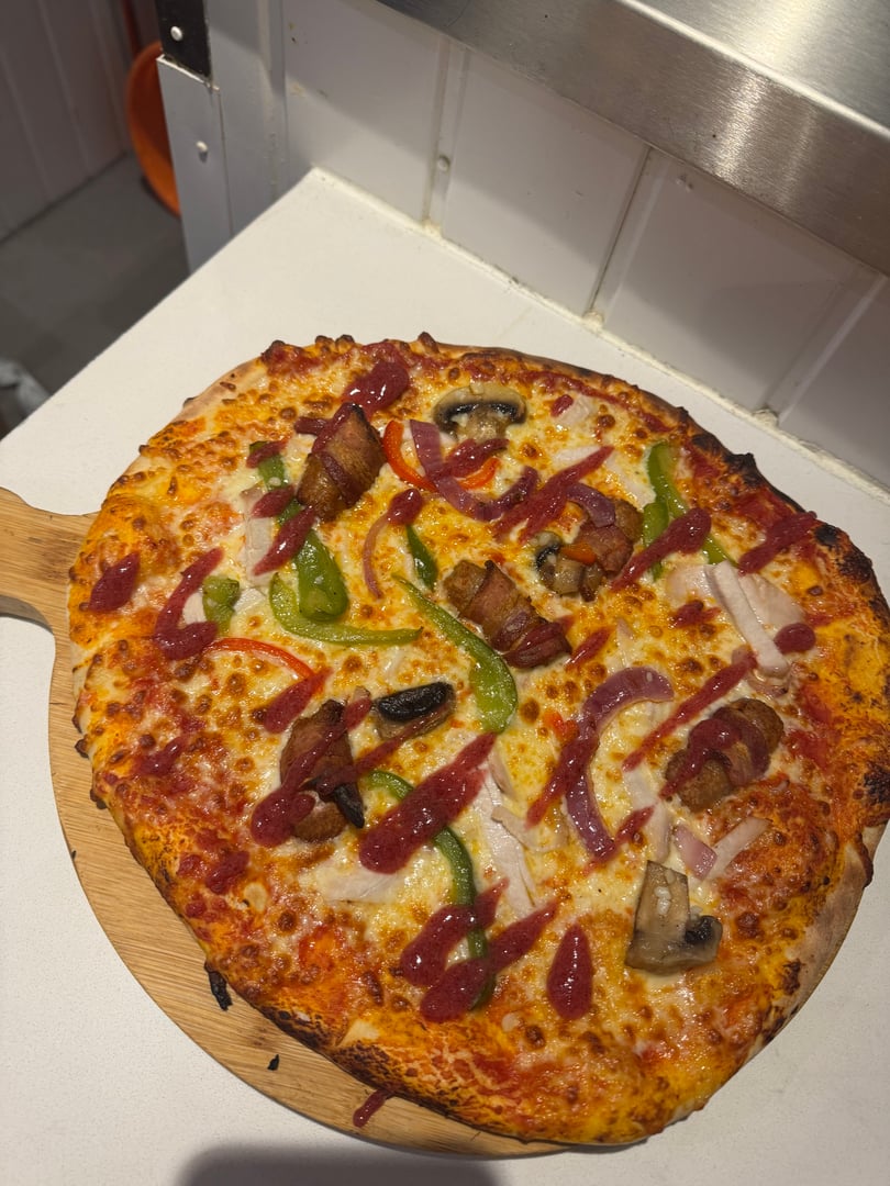 Pizza with bacon, green peppers, onions, mushrooms, and drizzled sauce on a wooden board.