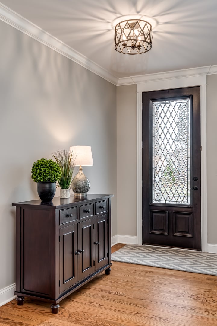 An entryway to a large home with plants and a lamp sitting on top of a cabinet with the glass front door behind.