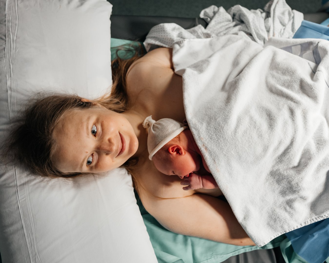 A mother smiles joyfully as she holds her newborn baby on her chest for skin-to-skin contact after childbirth, wrapped in a warm blanket in a hospital setting.