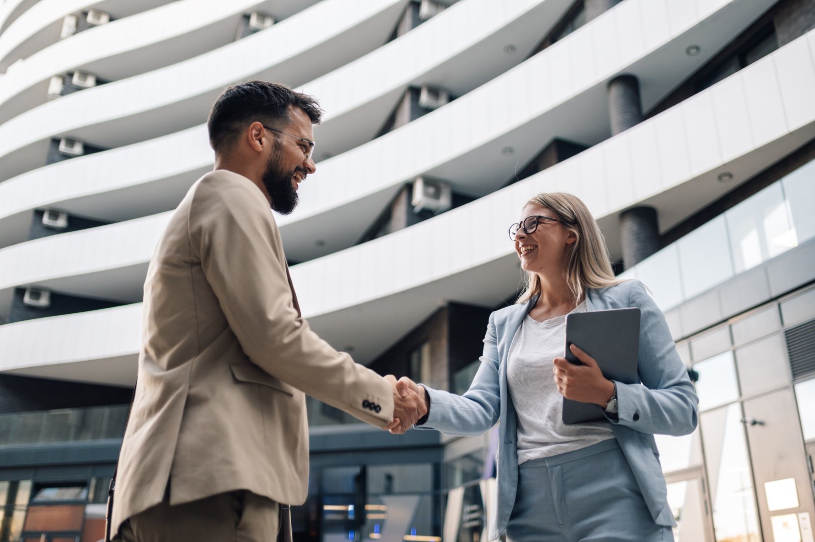 Two business partners happily shaking hands in front of office building, showcasing teamwork and collaboration