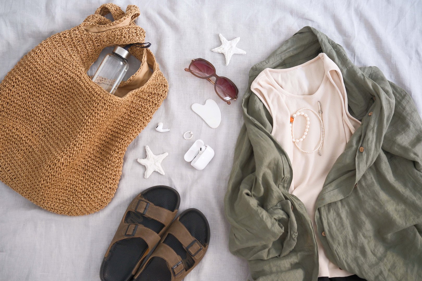 A trendy summer outfit is beautifully laid out along with chic accessories, making it perfect for a beach day - linen shirt, tee-shirt, straw bag, spanking, glasses.