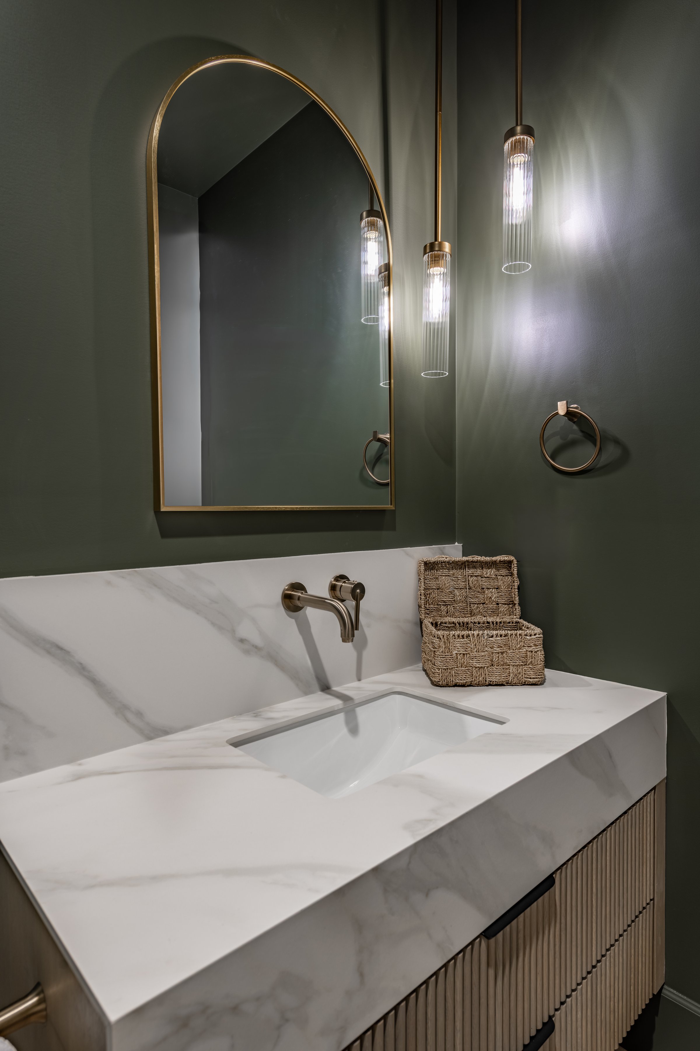 powder room interior with dark green painted walls pendant lighting hanging down large mirror with rounded top white marble countertop bronze gold faucet small basket on the counter top next to the sink
