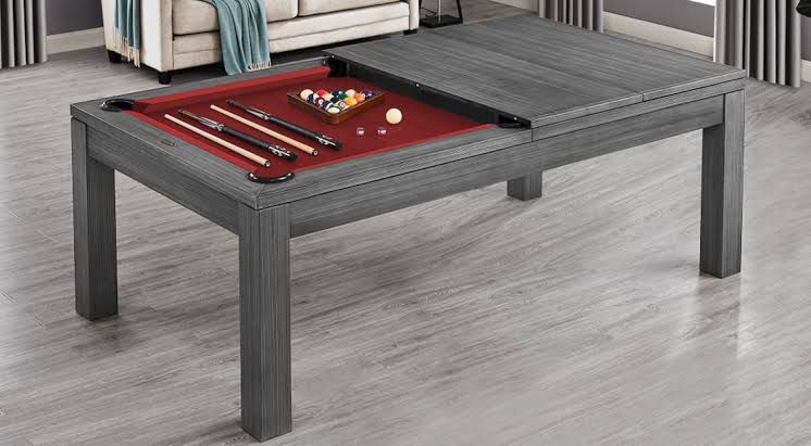 A gray billiard table with red felt, pool balls, and cues on top, set in a modern room with hardwood flooring.