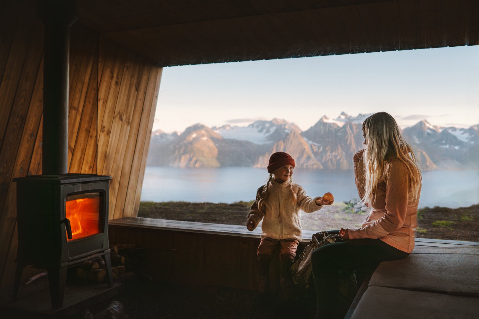 Family mother and child relaxing in cozy mountain hut with fireplace and panoramic window with mountains view, parent traveling with child in Norway autumn vacations trip, mom and kid eating snacks