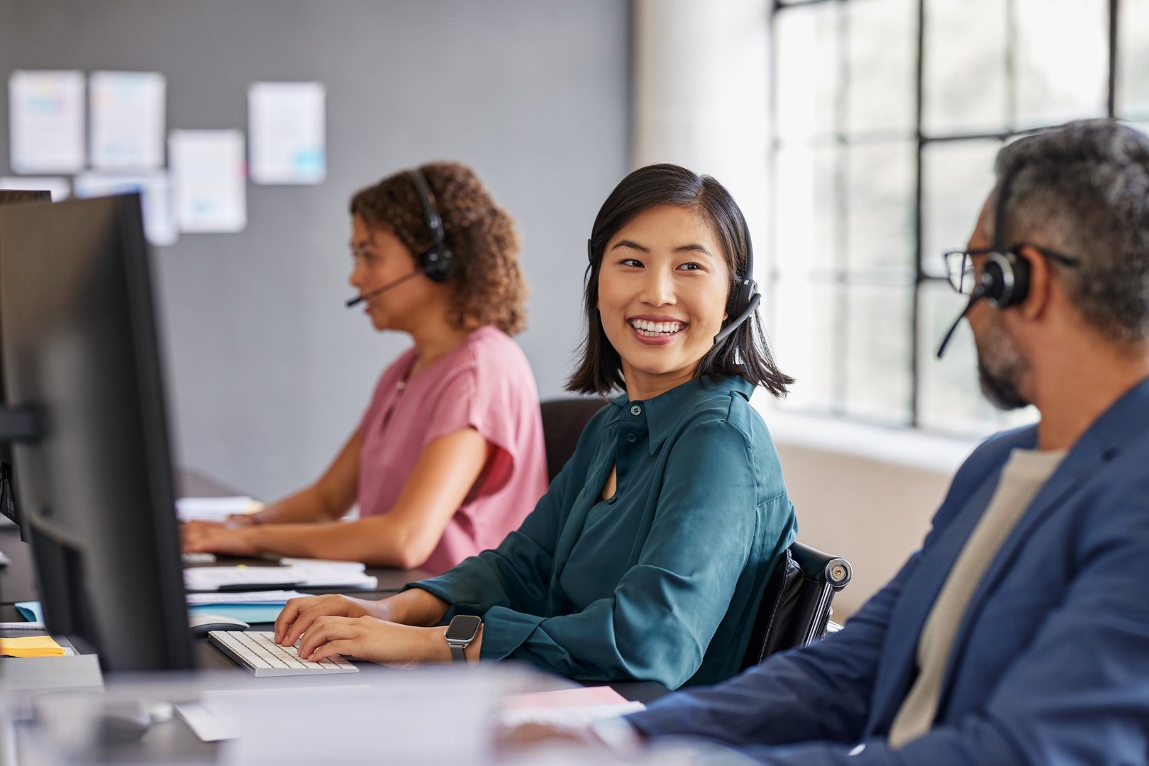 Happy customer service representative interacting with colleagues in a call center. Call center agent smiling while collaborating with support team. Young asian woman with headset talking to her supervisor while working.