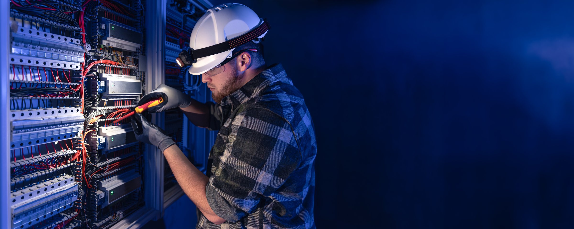 Male electrician working in switchboard with electrical connecting cable. Electrical engineer in special clothes with flashlight on helmet in dark room with emergency lights. Space for text.