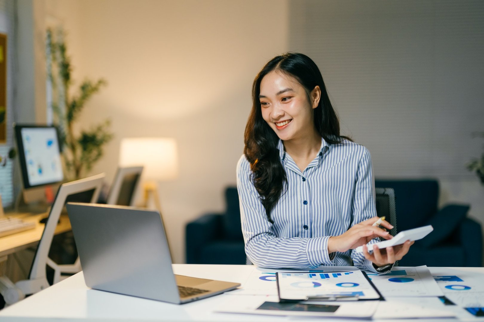 Young businesswoman working diligently at her desk in a modern office, using a calculator and laptop to analyze financial data during the night, focused on achieving her goals