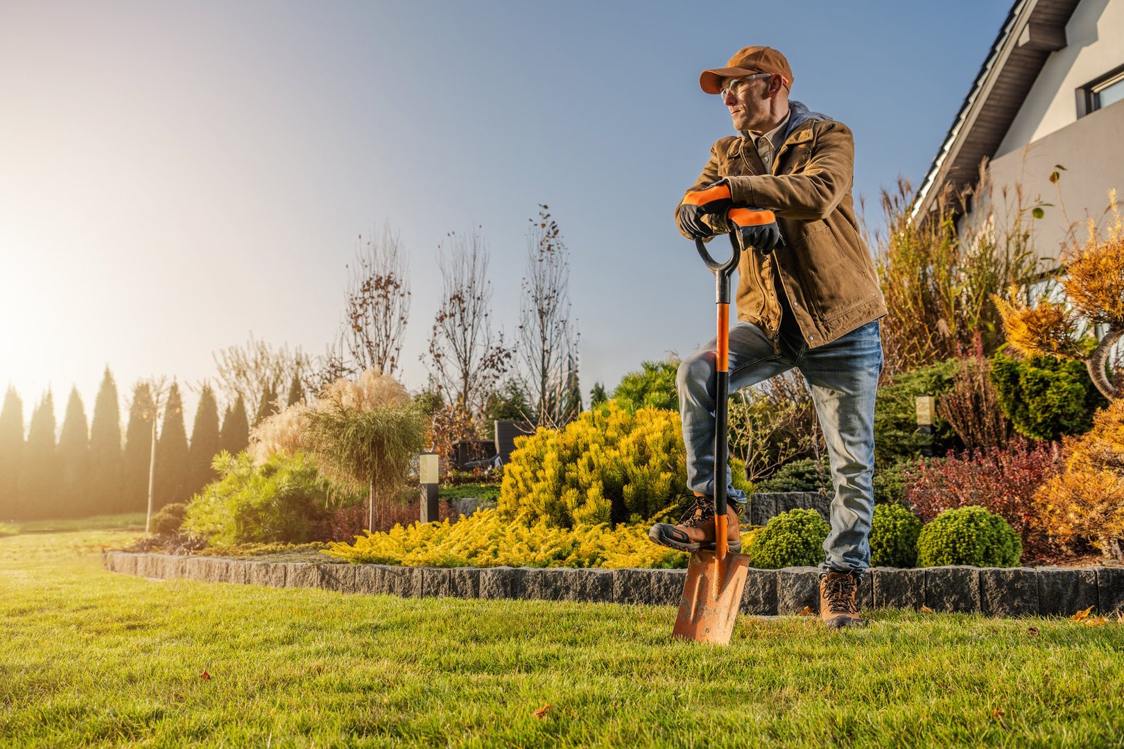 A man stands confidently with a shovel in the garden, enjoying the beautiful sunset. The vibrant landscape features well-kept plants and greenery.